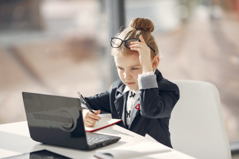 Młoda dziewczynka w garniturze siedzi przed laptopem z zamyśloną miną, prawą ręką trzyma okulary, lewą dłonią się podpiera, a w dłoni prawej trzyma długopis nad otwartym notesem.