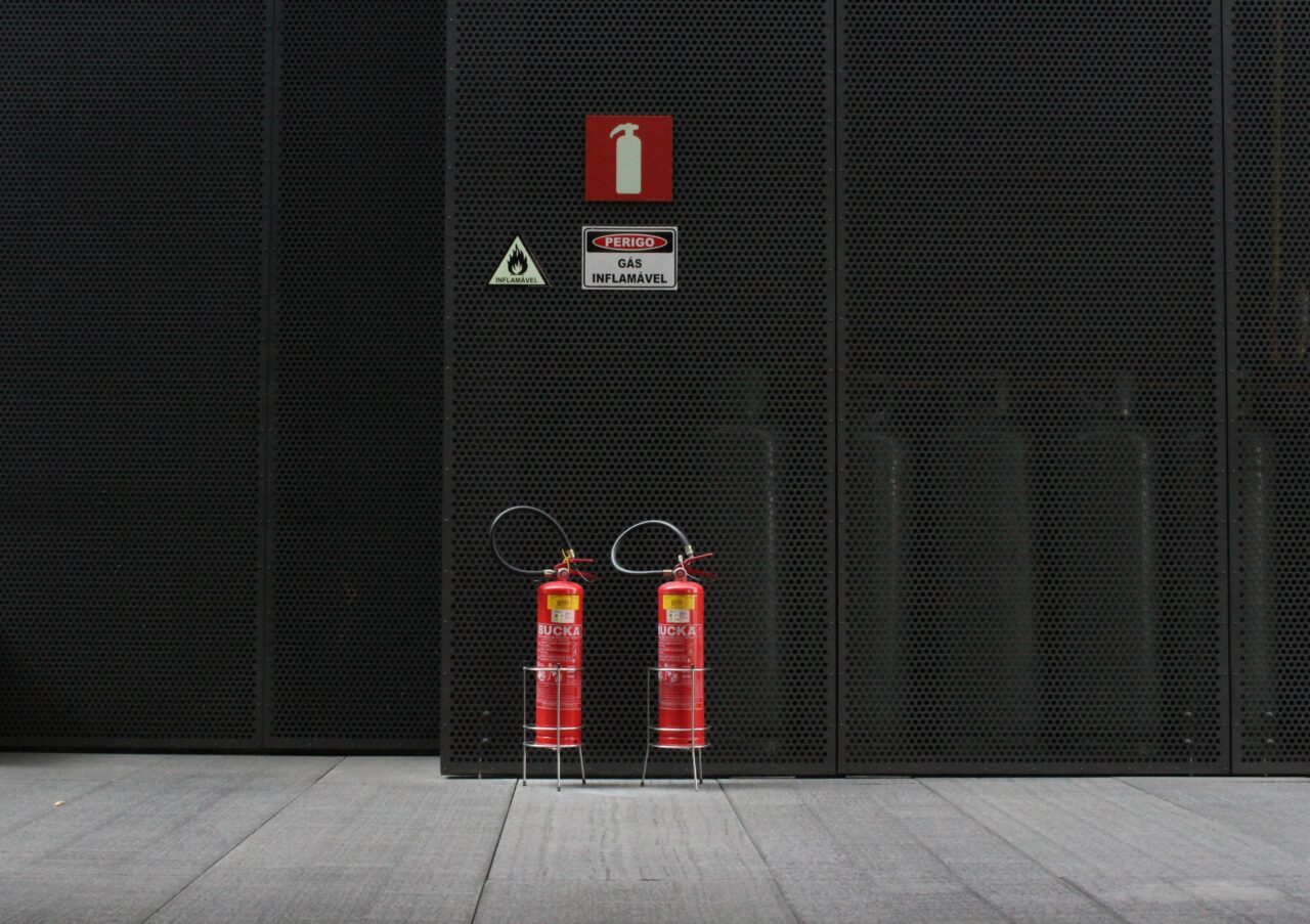 Scam telefoniczny. Dwa czerwone gaśnice stoją przed metalową siatką, na której umieszczone są znaki ostrzegawcze o przeciwpożarowym wyposażeniu i gazie łatwopalnym.