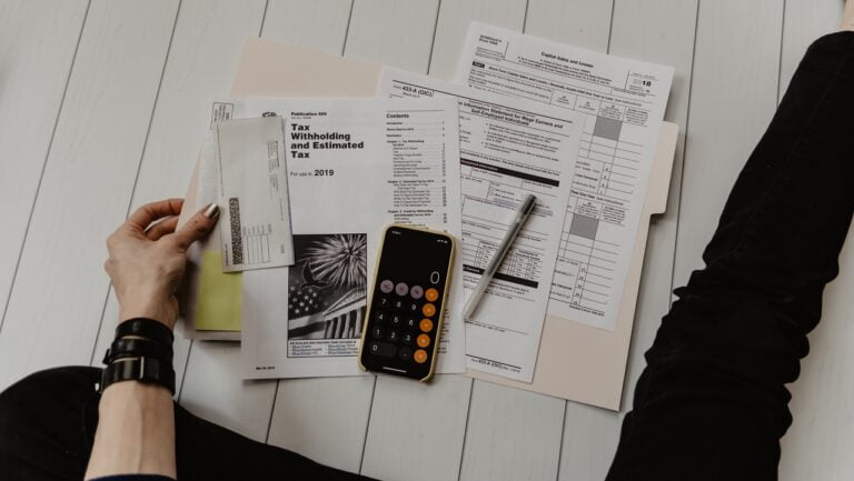 Osoba przeglądająca dokumenty podatkowe i korzystająca z kalkulatora na smartfonie, na podłodze leżą różne formularze podatkowe i długopis.