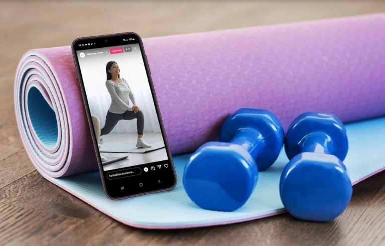 Smartfon leżący na skręconej maty do jogi wyświetla wideo z ćwiczącą kobietą, obok znajdują się dwa niebieskie hantle.