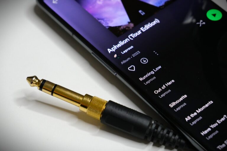 Złoty wtyk jack 6,3 mm leżący obok smartfona wyświetlającego ekran aplikacji Spotify z listą utworów z albumu "Aphelion (Tour Edition)" zespołu Leprous.