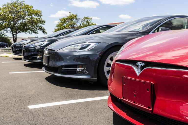 Samochody elektryczne marki Tesla zaparkowane w rzędzie na parkingu, z widocznymi charakterystycznymi logo przedstawiającymi literę "T" na czerwonobrązowych i srebrnoszarych przednich maskach.