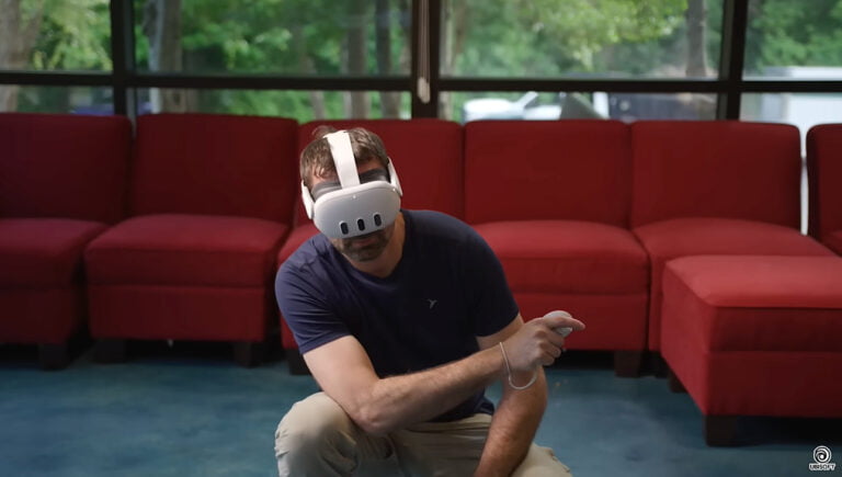 Kucający mężczyzna w pokoju z goglami VR założonymi na głowę. W tle czerwona kanapa