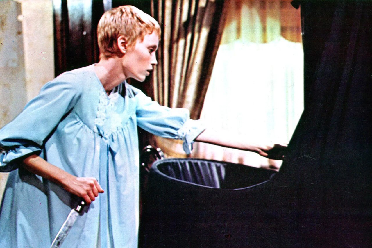 Kadr z filmu Dziecko Rosemary. Młoda kobieta o krótkich włosach ubrana w błękitną nocną koszulę trzyma miecz, delikatnie odsłaniając zasłonę w zaciemnionym pomieszczeniu.