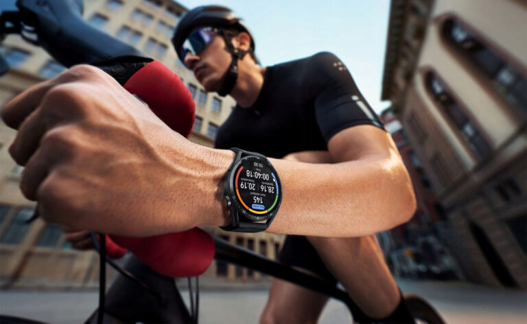 Kolarz w kasku i okularach przeciwsłonecznych trzyma kierownicę roweru, na jego nadgarstku widoczny smartwatch, w tle miejska zabudowa.
