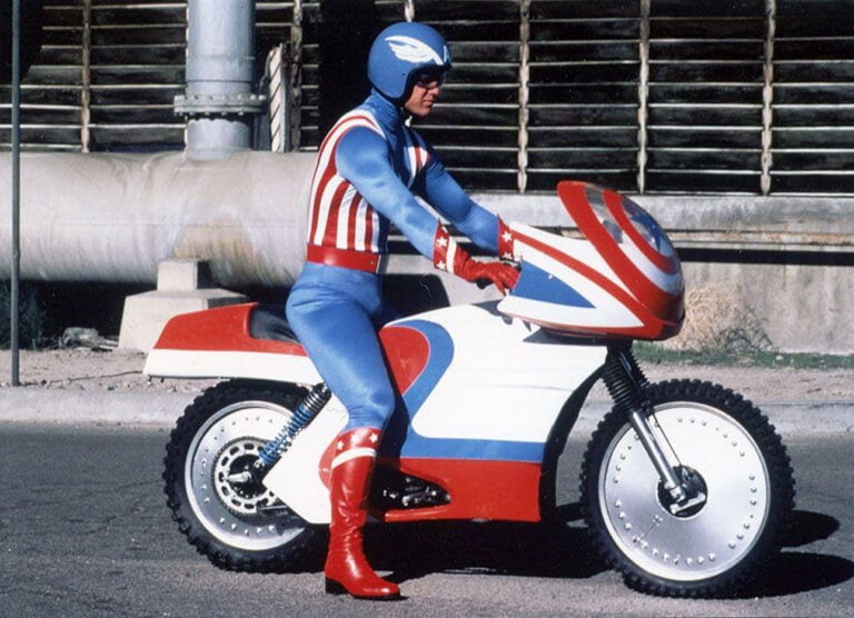 Reb Brown jako Kapitan Ameryka siedzący na motocyklu. Ubrany w niebiesko czerwony kostium superbohatera. Motocykl w tych samych kolorach