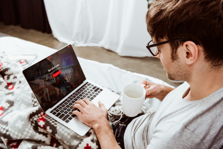 Mężczyzna w okularach korzystający z laptopa z wyświetlonym interfejsem platformy streamingowej Netflix, trzymający kubek, siedzący na kanapie przykrytej kocem w jodełkę.