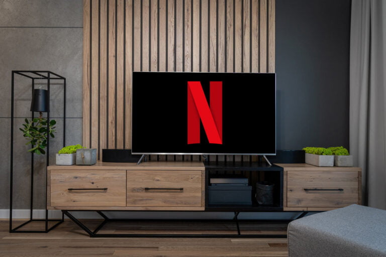 w centrum zdjęcia znajduje się telewizor z logo Netflix na ekranie. Urządzenie stoi na komodzie z drewnianymi szufladami. W tle na ścianie widać drewiane deski