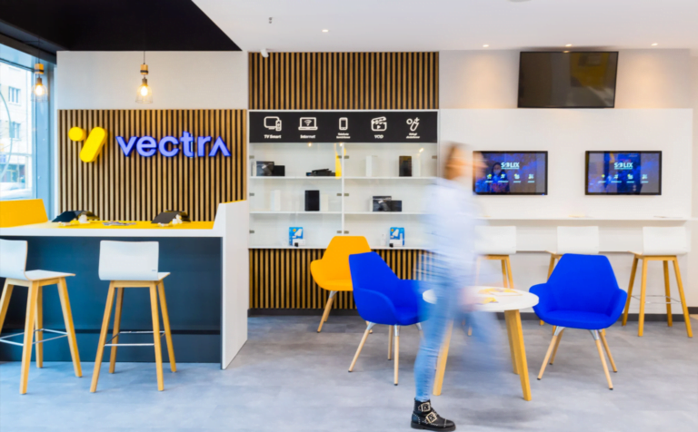Jasne, nowoczesne wnętrze punktu obsługi klienta Vectra, z widocznym logo firmy, stolikami i krzesłami barowymi oraz rozmytym zdjęciem przechodzącej osoby.