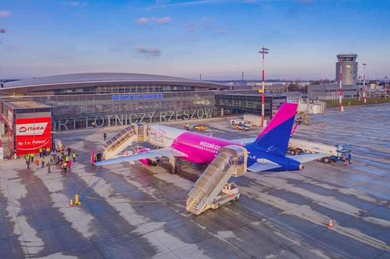 Samolot pasażerski Wizz Air stoi przy bramce na Lotnisku Rzeszów-Jasionka z pasażerami wsiadającymi po schodach; w tle budynek terminala i wieża kontroli lotów.