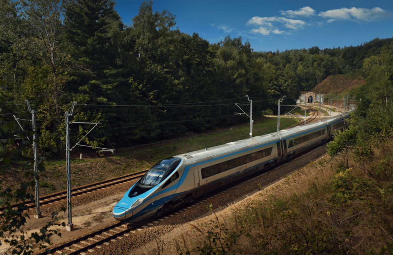 Pociąg pendolino na torach w zalesionym terenie, w tle tunel.