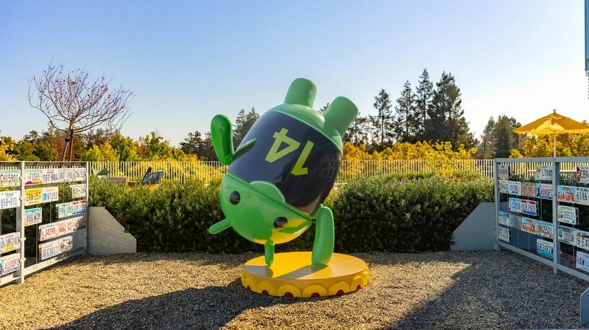 Duży, zielony pomnik Androida na zewnątrz z czarną twarzą i symbolem błyskawicy, otoczony tabliczkami rejestracyjnymi i zielenią w tle.