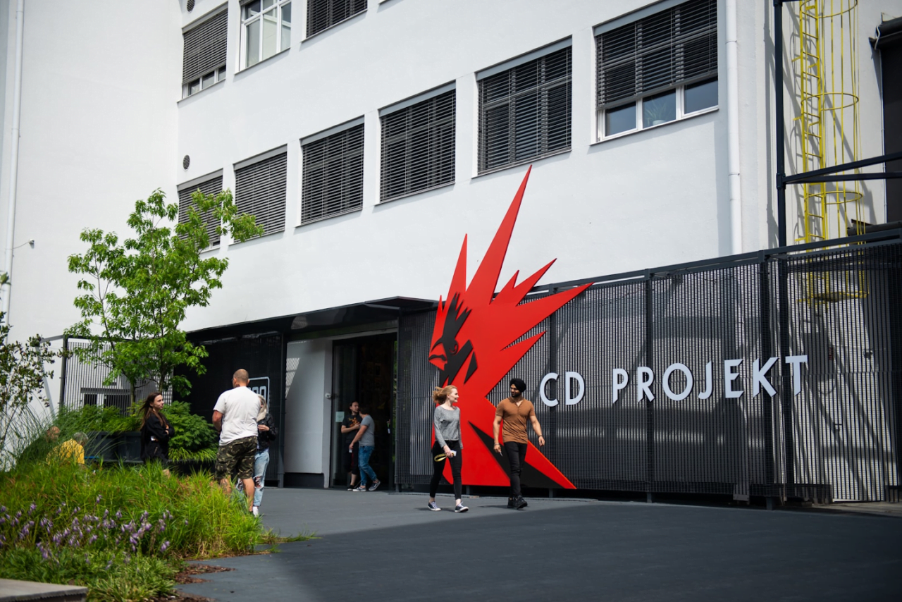 Wejście do siedziby CD Projekt RED z dużym czerwonym logo w kształcie ptaka na ścianie budynku, ludzie przechodzący obok.