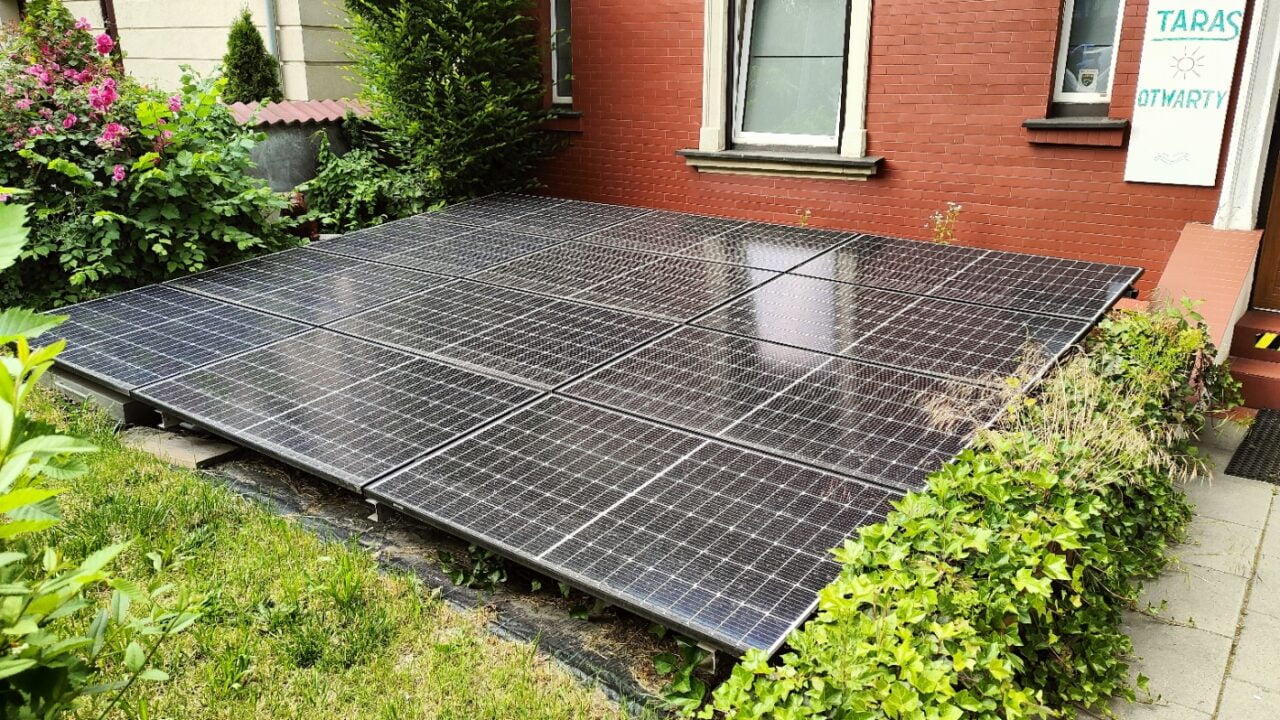 Panele słoneczne, wykrywalne przez Google Solar API zamontowane na ziemi przed budynkiem z czerwoną ceglaną elewacją, otoczone roślinnością.