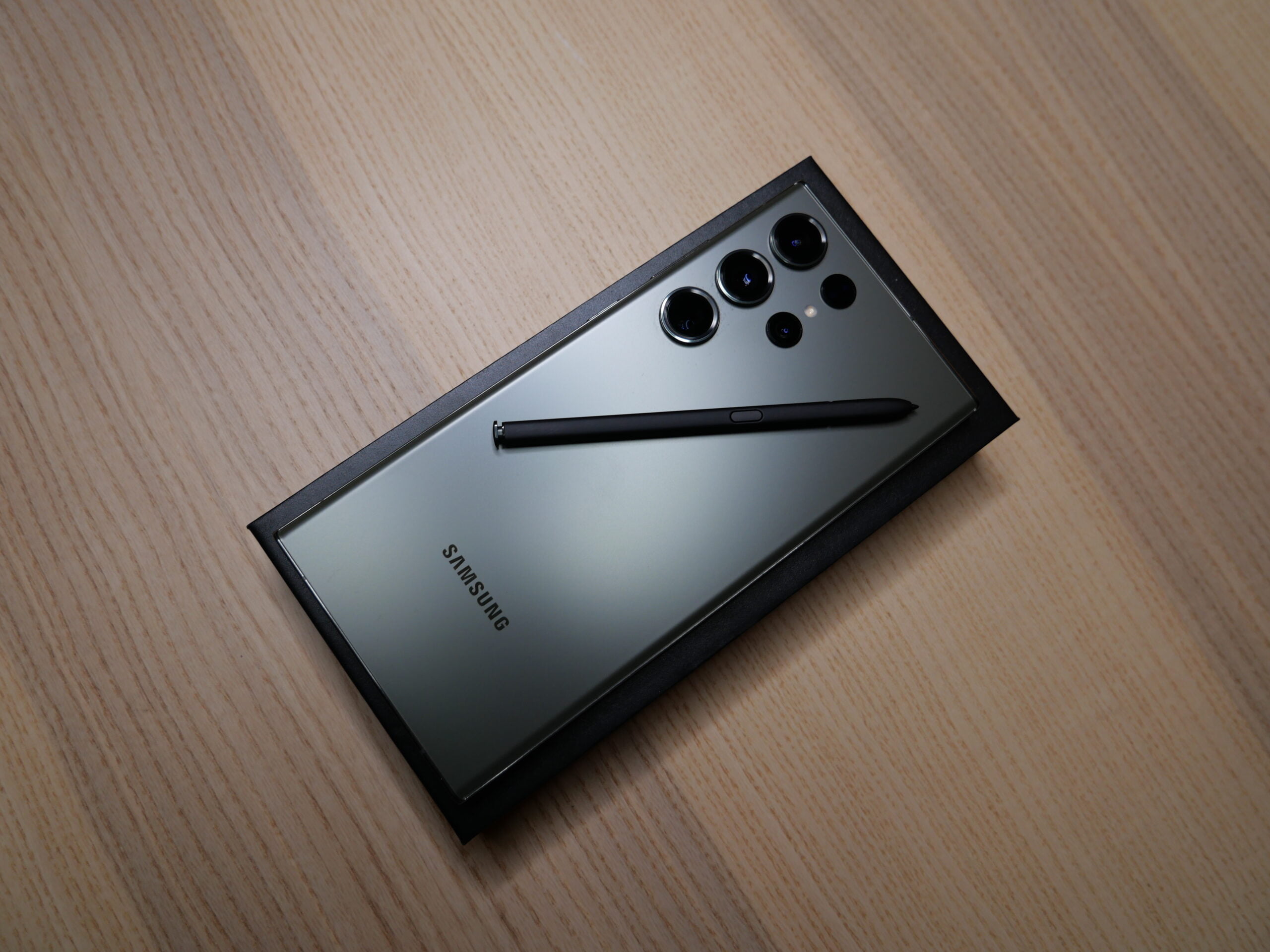 Czarny smartfon Samsung Galaxy Note z rysikiem S Pen położonym na jego tylnej części, leżący na drewnianym biurku.