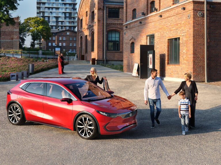 Czterech ludzi w miejskiej scenerii przy czerwonym samochodzie marki Izera, dwoje dorosłych i dziecko idą trzymając się za ręce, a kobieta wychodzi z pojazdu.
