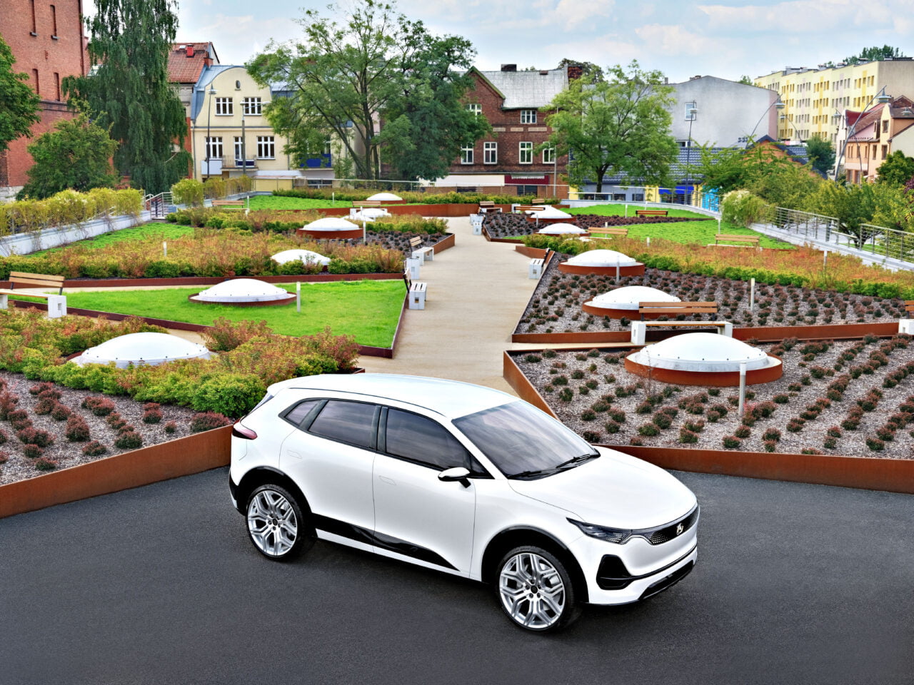 Biały SUV Izera ujęty z dalszej perspektywy bokiem na tle parku z roślinami.