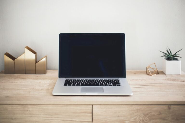 Otwarty laptop stojący na drewnianym meblu. W tle złote ozdoby, roślina i jasna ściana