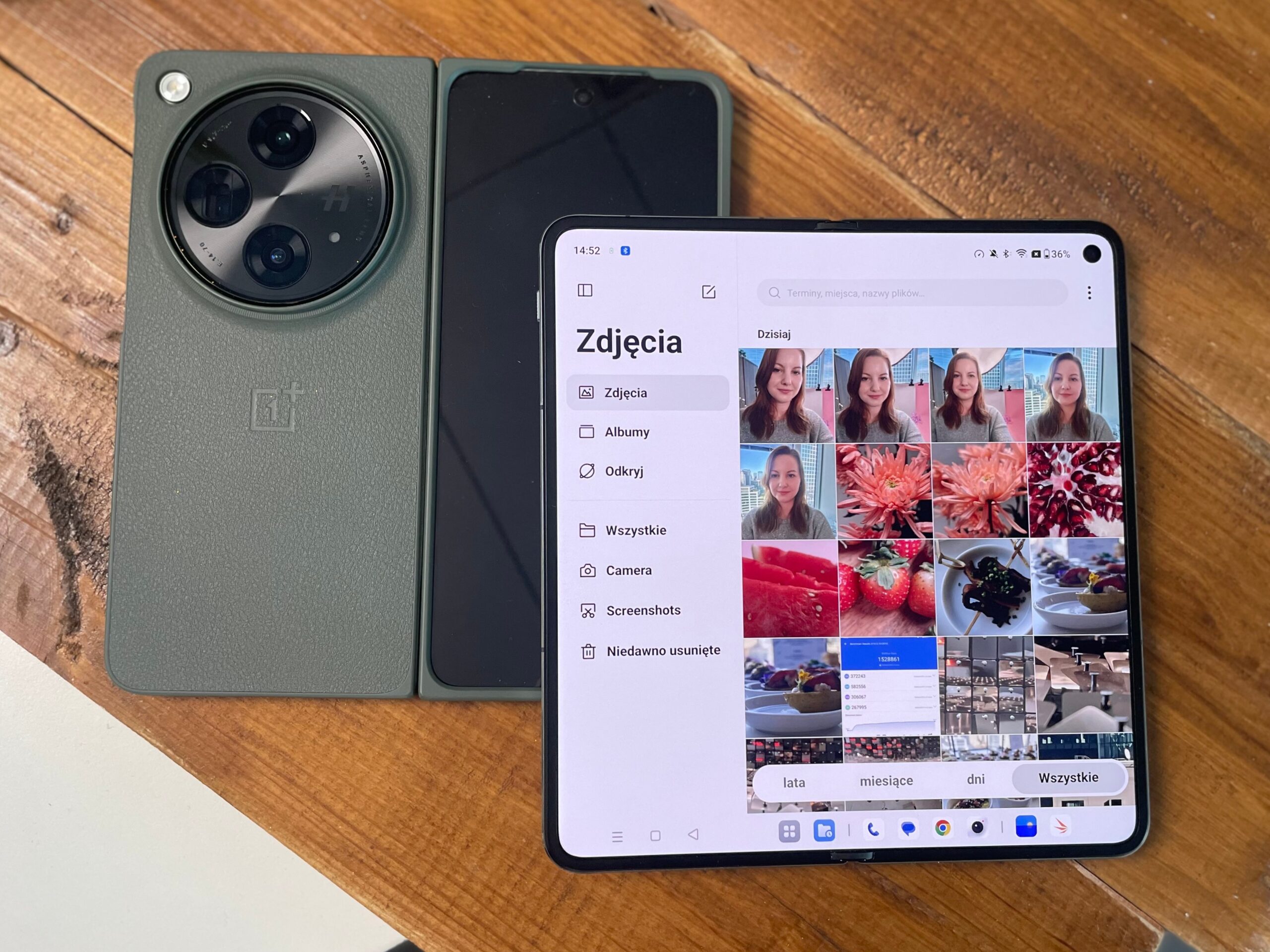 Zdjęcie przedstawia zielony smartfon z aparatem o trzech obiektywach i OnePlus Open z otwartą aplikacją galerii wyświetlającą zdjęcia, oba urządzenia leżą na drewnianym blacie.
