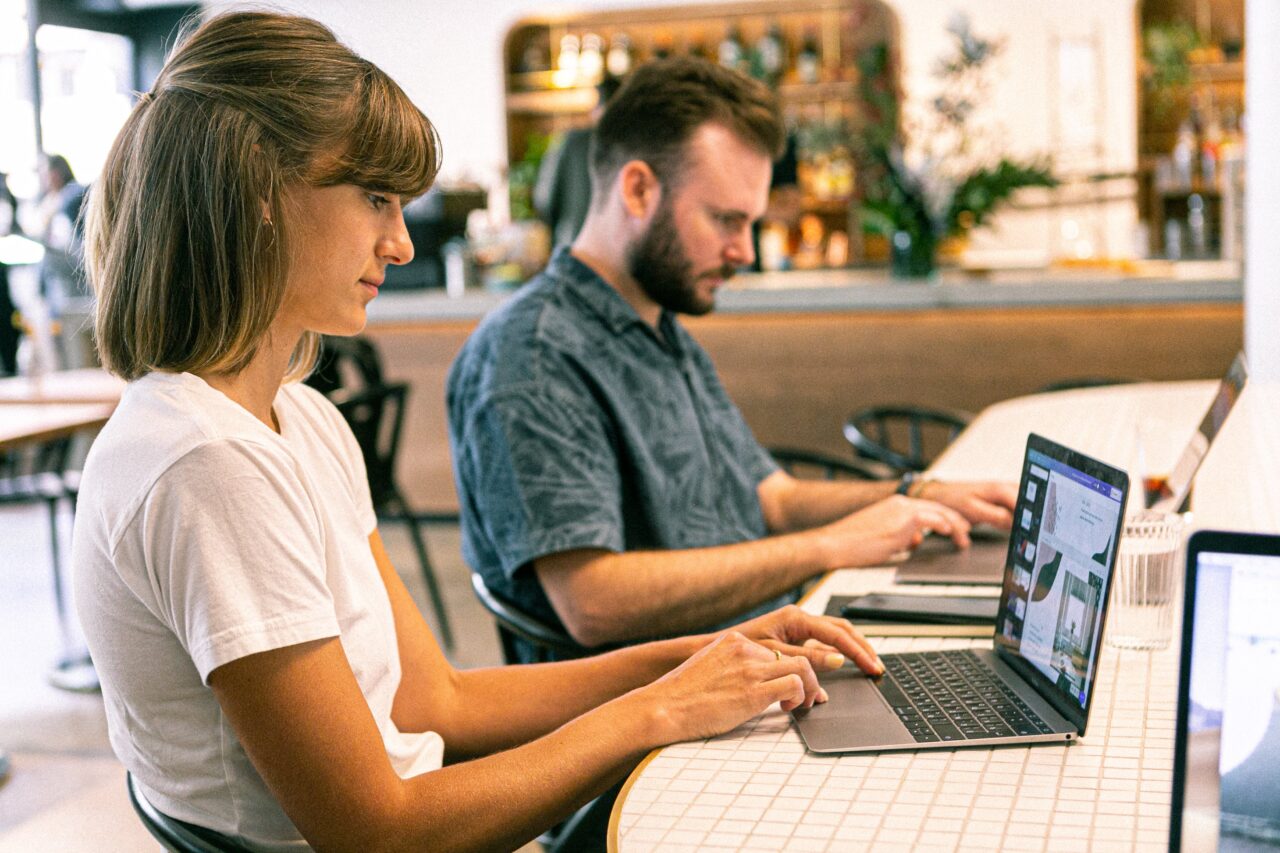 Kobieta i mężczyzna sfotografowani z boku pracują na laptopach w kawiarni.