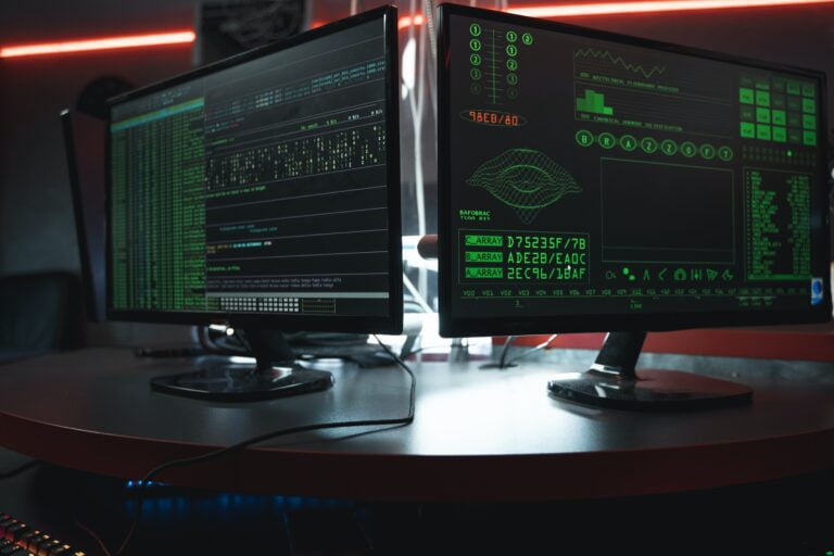 Dwa monitory komputerowe z włączonym oprogramowaniem do programowania lub analizy danych, postawione na biurku w słabo oświetlonym pomieszczeniu z czerwonym światłem w tle.