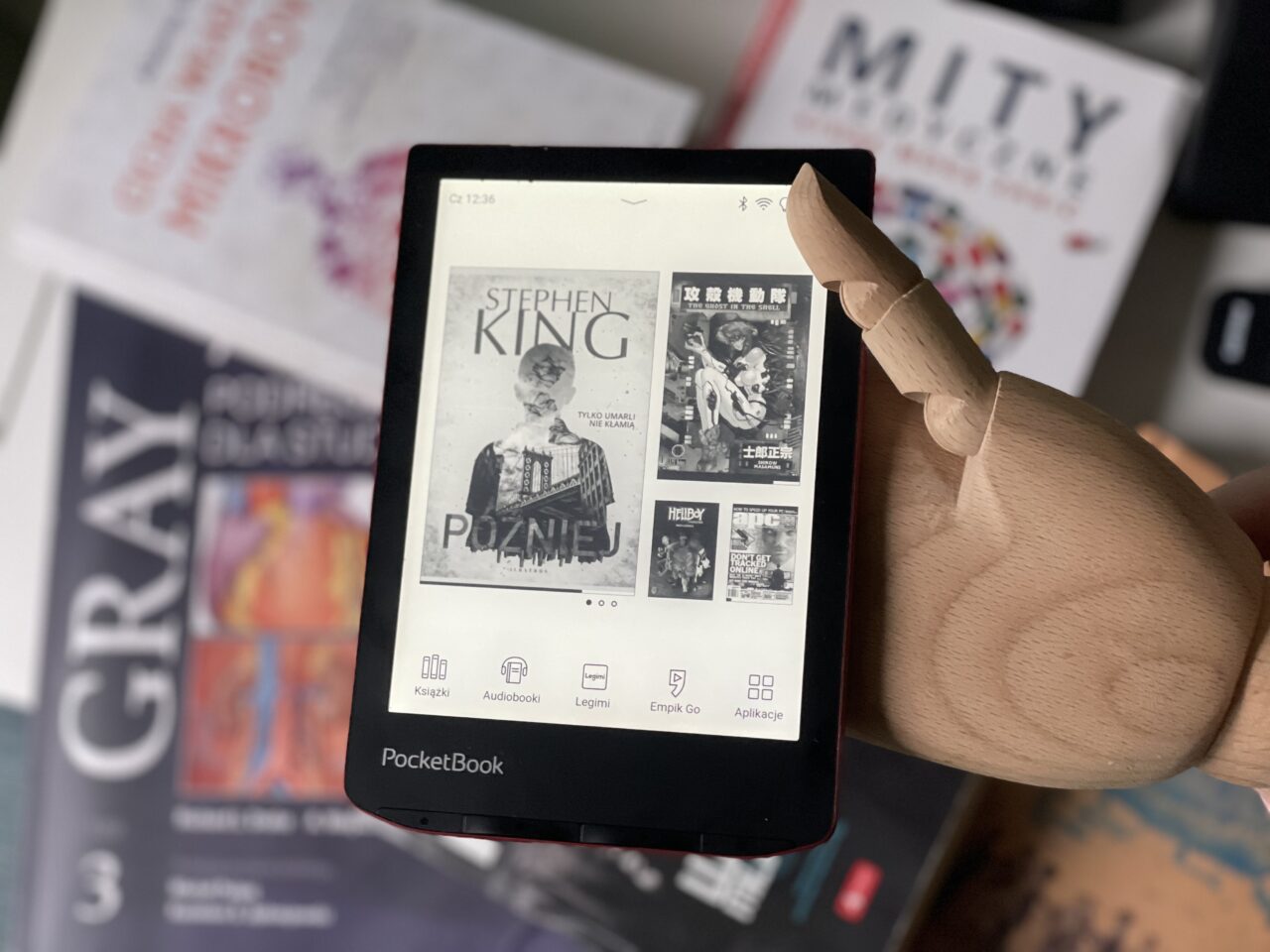 Czytak elektroniczny PocketBook trzymany przez drewnianą rękę, na ekranie widoczne okładki e-książek, w tym "Później" Stephena Kinga, otoczony przez inne przedmioty, takie jak książki i czasopisma rozłożone w nieładzie.