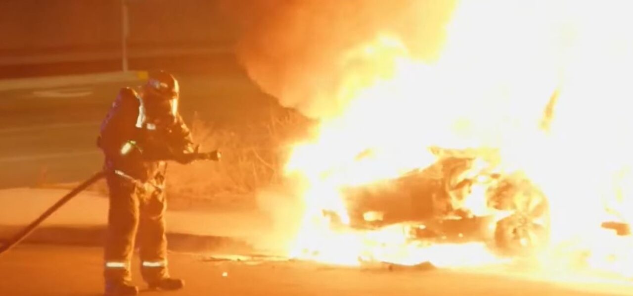 Strażak gaszący płonący samochód elektryczny w nocy.