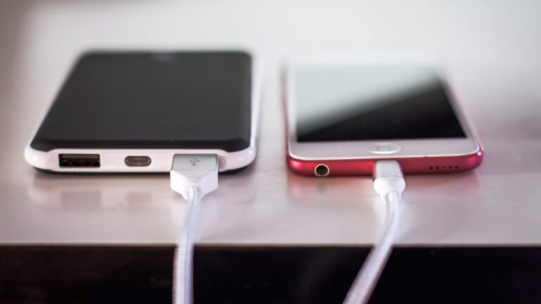Dwa smartfony leżące obok siebie na biurku, każdy podłączony do białego kabla ładującego; po lewej czarny telefon z portem micro-USB, po prawej czerwono-biały telefon z portem Lightning.