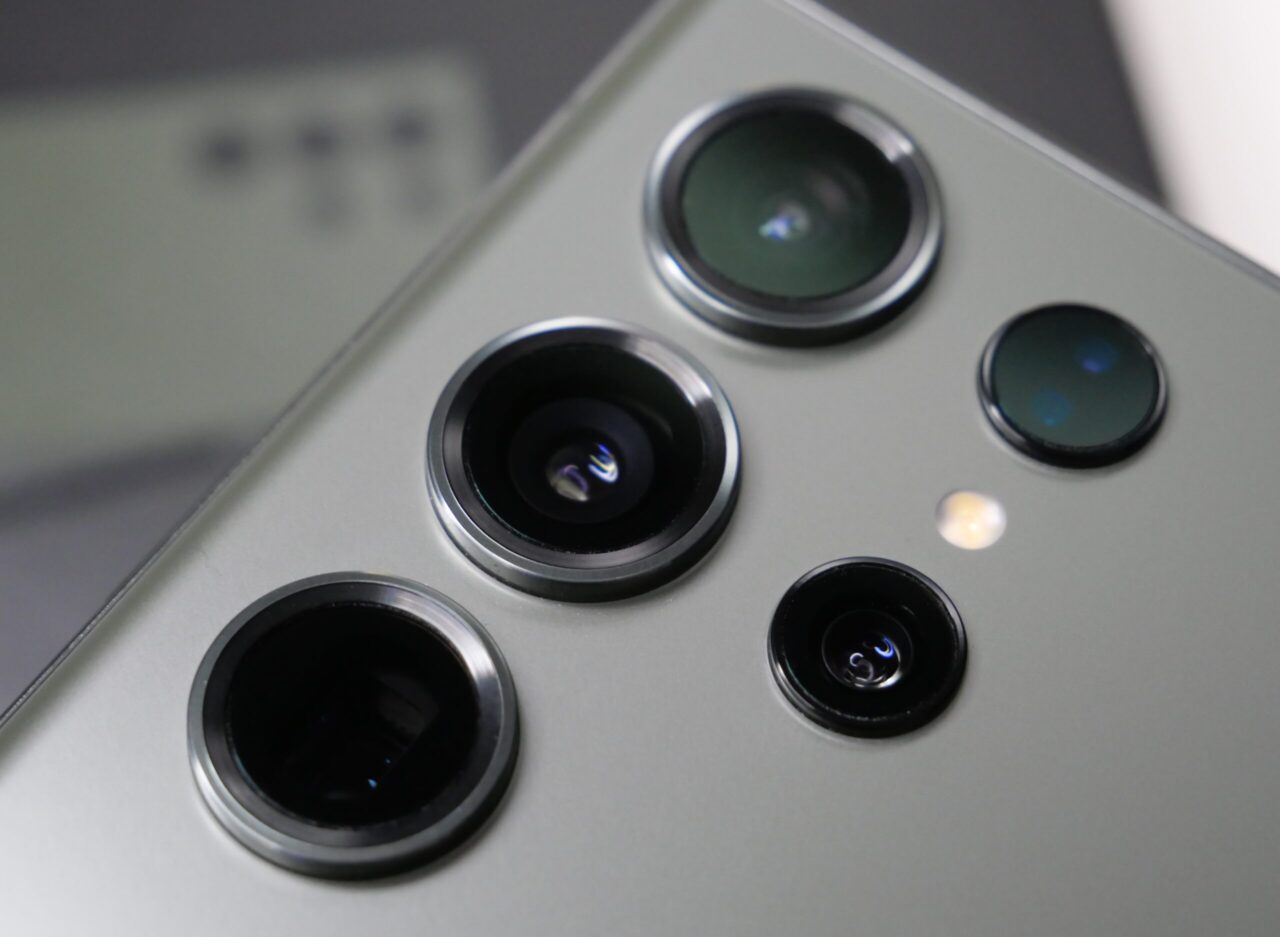 Zbliżenie na aparat fotograficzny smartfona Galaxy S23 Ultra z trzema obiektywami i czujnikiem światła, na szarym tle.