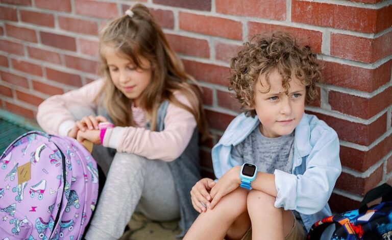 Dwoje dzieci siedzi na ziemi oparte o ceglaną ścianę; dziewczynka po lewej strony z zegarkiem na nadgarstku patrzy na swój plecak, a chłopiec po prawej stronie z zegarkiem na nadgarstku wygląda na zamyślonego.