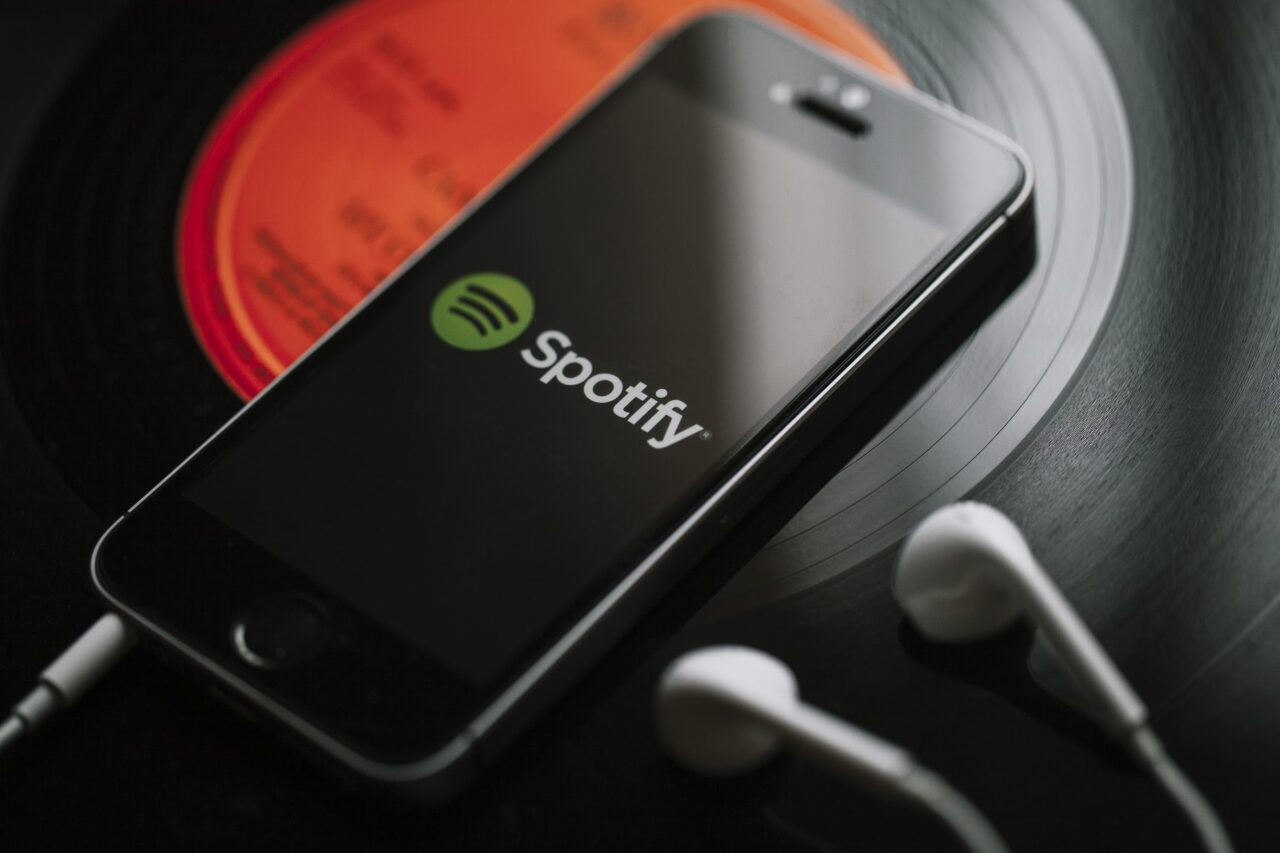 Smartfon z wyświetlonym logo Spotify na czarnym tle leżący na płycie winylowej. Do smartfona podpięte są białe słuchawki douszne.