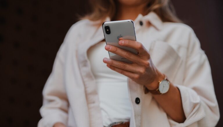 Kobieta w białej koszuli trzyma w dłoni srebrny smartfon, na jej nadgarstku widoczny jest złoty zegarek.