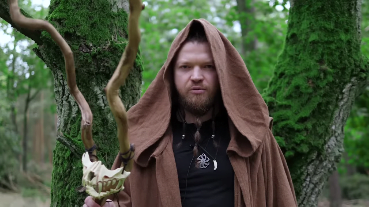 Sylwester Wardęga mówi „pas”. Mężczyzna w brązowym płaszczu z kapturem, stojący w lesie, trzymający kostur z czaszką jelenia.