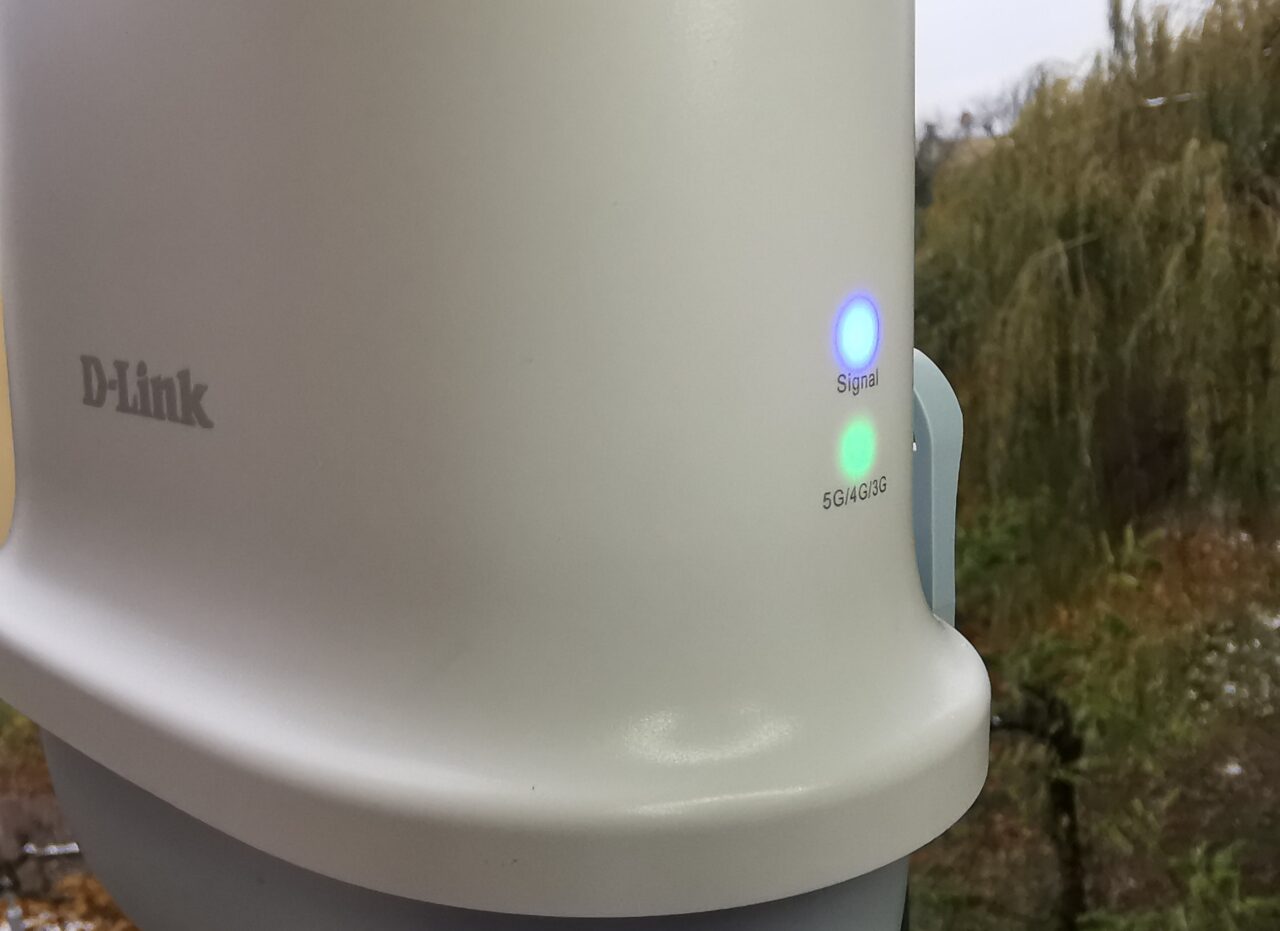 Część białego urządzenia sieciowego marki D-Link z włączonymi kolorowymi diodami sygnalizującymi siłę sygnału oraz obsługiwane pasma 5G/4G/3G, na nieostro widoczne drzewa w tle.
