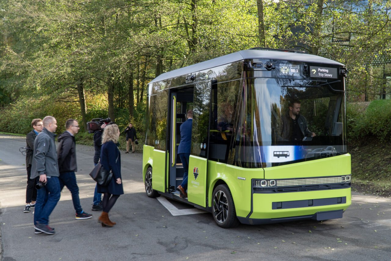 Na zdjęciu znajduje się autonomiczny bus w fazie testów z Katowic. Pojazd stoi w miejscu, ma zieloną karoserię, wchodzą do niego grupki ludzi, a w tle znajduje się leśna droga.
