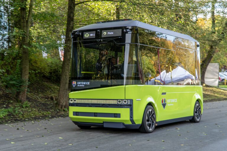Na zdjęciu znajduje się autonomiczny bus w fazie testów z Katowic. Pojazd jest w ruchu, ma zieloną karoserię, a w tle znajduje się leśna droga.