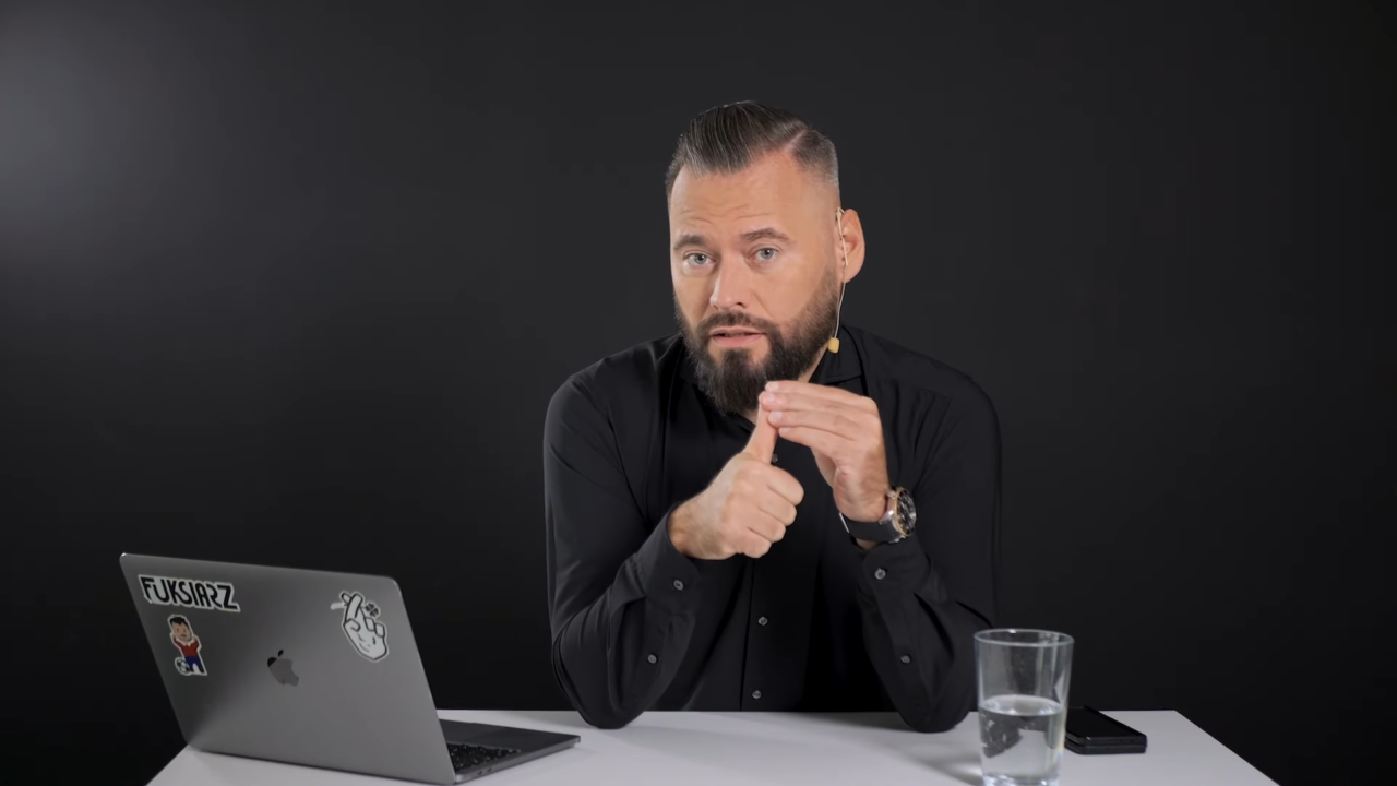Kanał Zero - Krzysztof Stanowski w czarnej koszuli siedzi przy biurku z laptopem i szklanką wody, wskazuje ręką, patrząc w kamerę na czarnym tle.