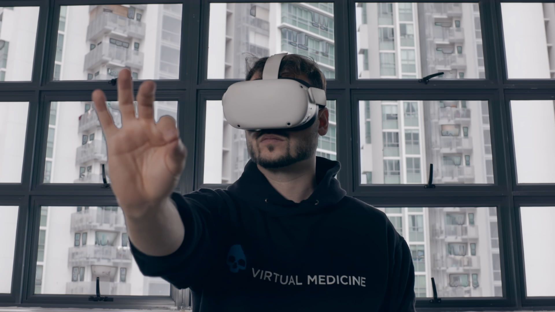 mężczyzna w goglach VR na głowie wyciąga przed siebie rękę. W tle widać okno oraz zabudowę miejską