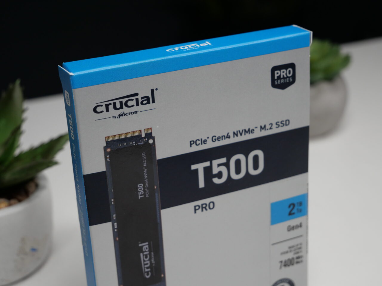 Niebiesko-białe opakowanie dysku SSD marki Crucial T500 Pro PCIe Gen4 NVMe M.2 o pojemności 2 TB, z widocznym zdjęciem produktu na froncie, stoi na stole obok doniczki z zieloną rośliną na tle rozmytego białego tła.