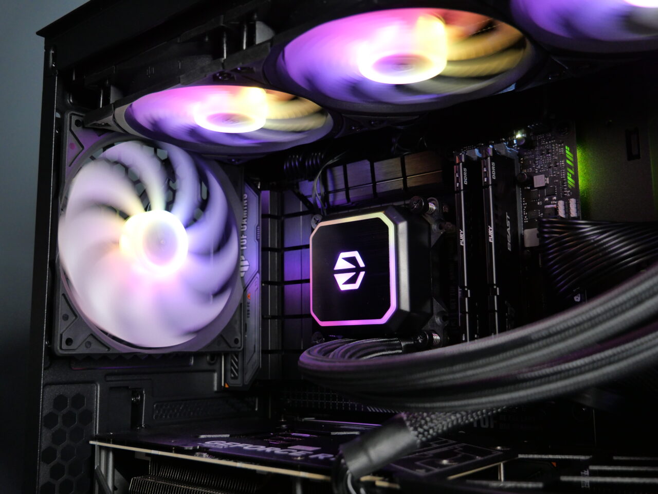 Wnętrze obudowy komputera z widocznymi elementami, takimi jak podświetlana na fioletowo płyta główna, trzy wentylatory z efektami RGB.