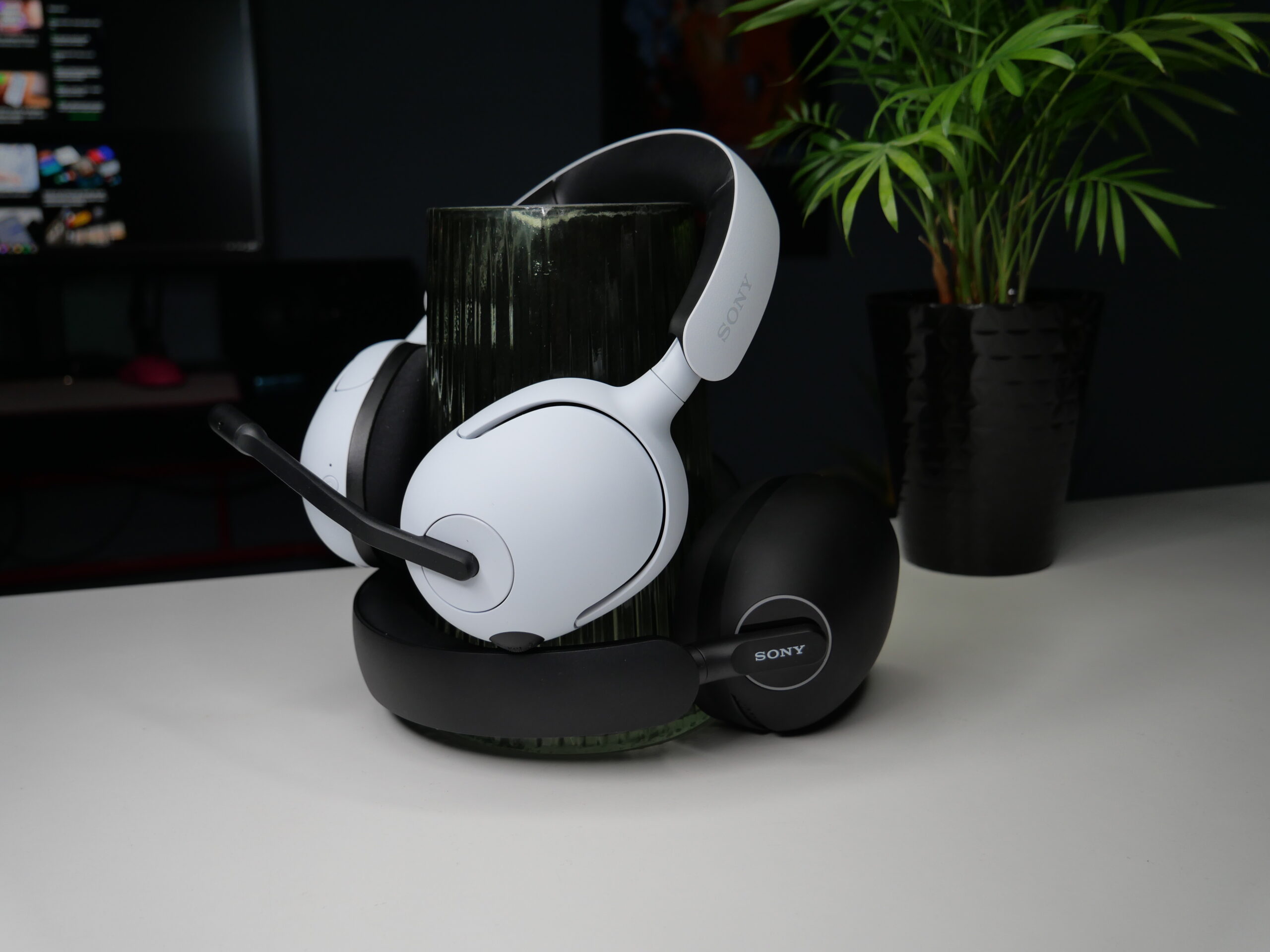 Białe słuchawki z mikrofonem marki Sony na stojaku, z roślinami w tle i monitorem w ciemnym pokoju.
