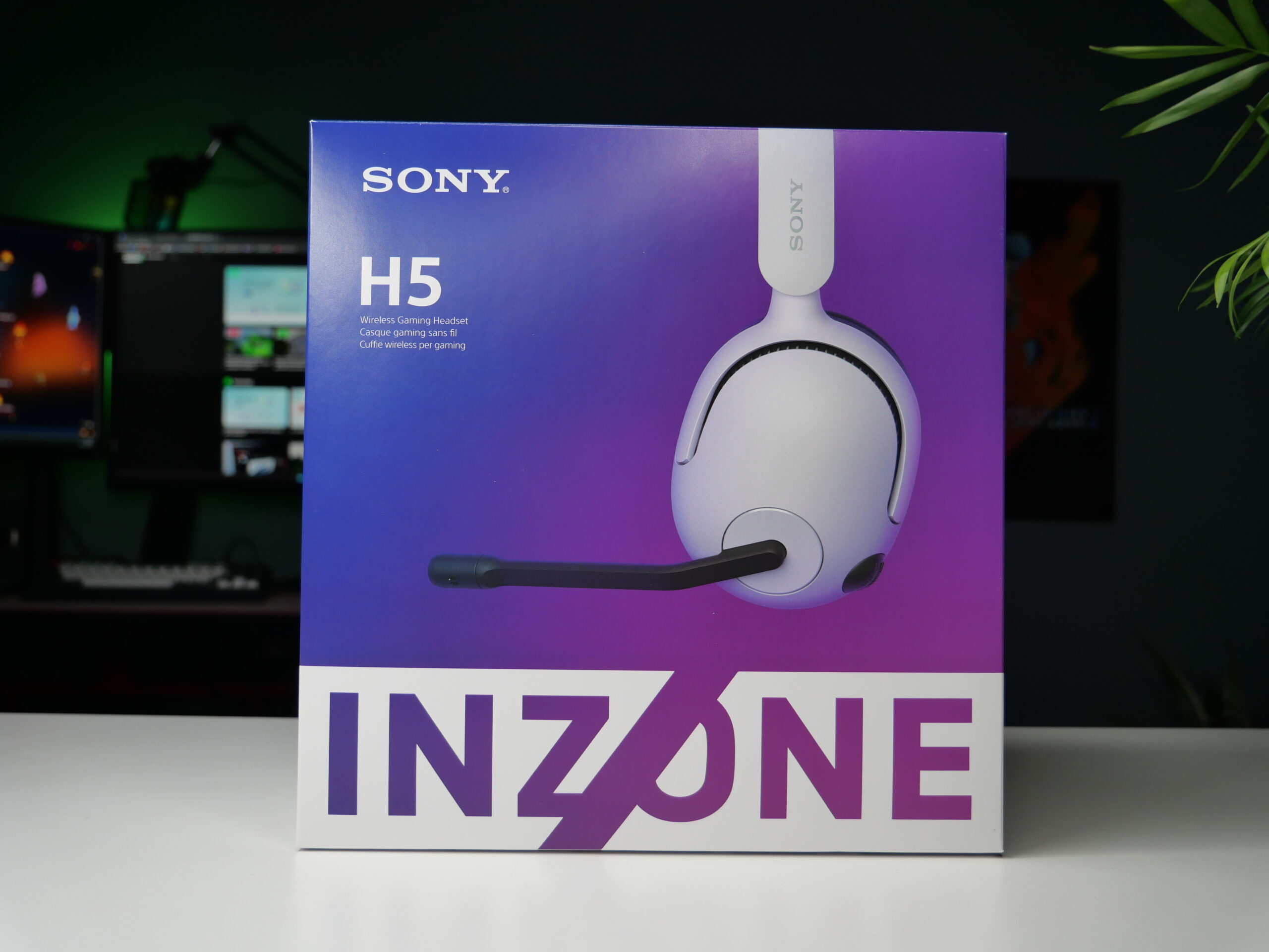 Opakowanie bezprzewodowych słuchawek gamingowych Sony INZONE H5 stoi na biurku, w tle widoczne są monitory komputerowe i część rośliny doniczkowej.