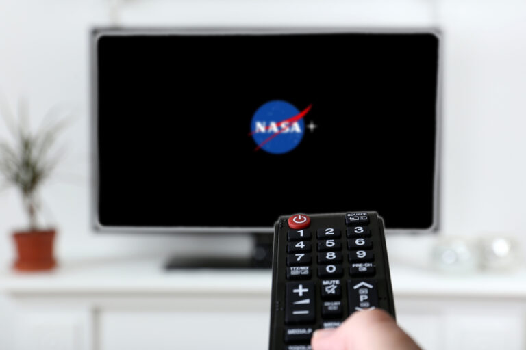 Telewizor powieszony na ścianie w salonie z logo NASA+ na ekranie. Na pierwszym planie fragment pilota i kawałek palca