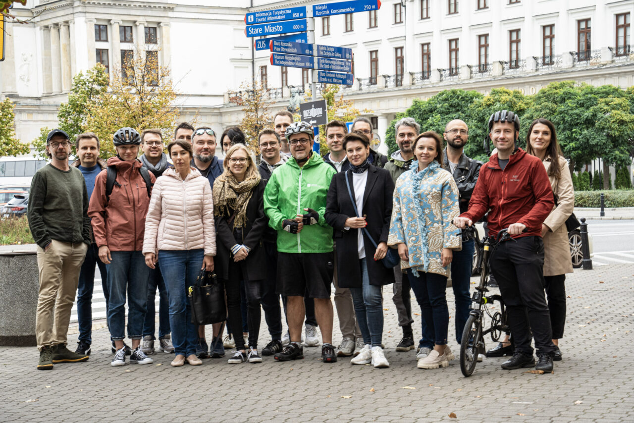 Grupa osób stojących na chodniku w miejskiej scenerii, z drzewami i budynkami w tle oraz znakami drogowymi wskazującymi kierunki do różnych atrakcji turystycznych. Elektryki i Wielki Test Mobilności Miejskiej 2023.