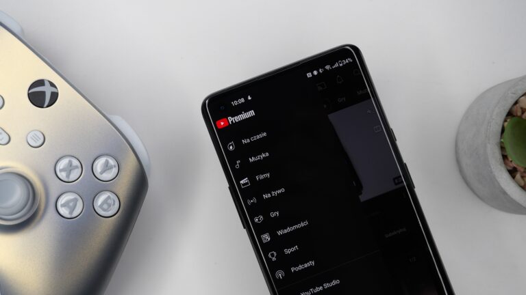 Srebrny kontroler do gier leży obok czarnego smartfona z otwartą aplikacją YouTube Premium i widocznym interfejsem menu. W tle doniczka z rośliną.