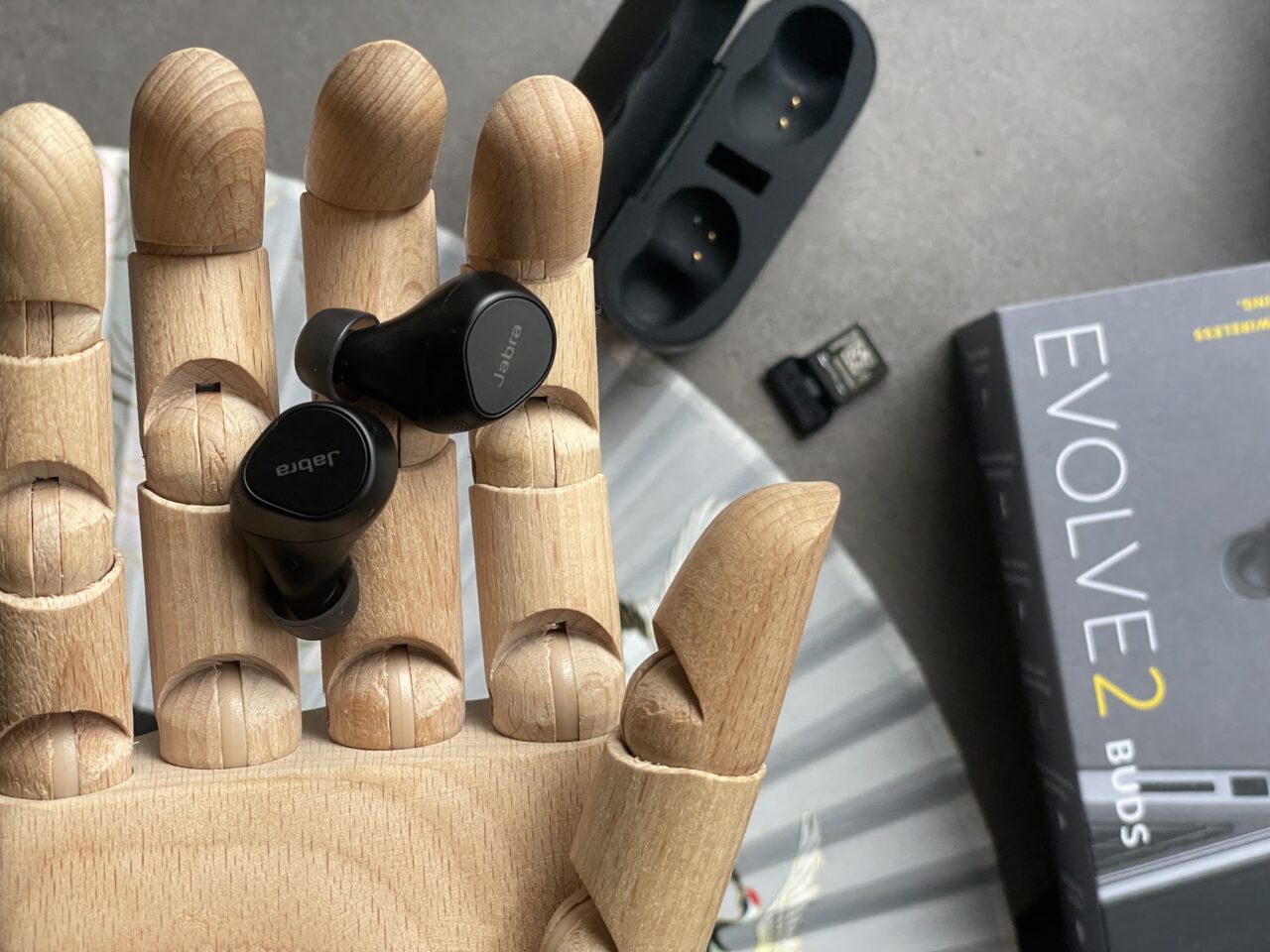 Drewniana ręka trzymająca czarne bezprzewodowe słuchawki douszne; w tle etui do ładowania słuchawek i pudełko z napisem "EVOLVE2 BUDS".