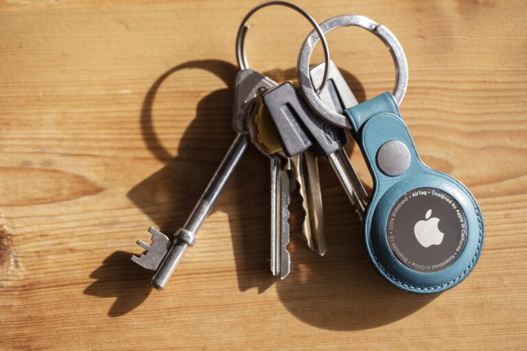Zdjęcie przedstawia zestaw kluczy leżących na drewnianej powierzchni, na które pada światło słoneczne tworząc ich cienie. Wśród kluczy znajduje się jeden o staromodnym kształcie. Klucze są połączone za pomocą metalowych kółek. Wyróżnia się brelok z niebieskim uchwytem i logo Apple, lokalizator AirTag