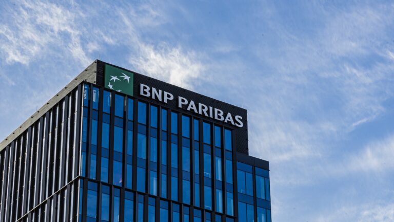 Widok na górne partie nowoczesnego budynku z logo BNP Paribas na szczycie na tle niebieskiego nieba z białymi chmurami.