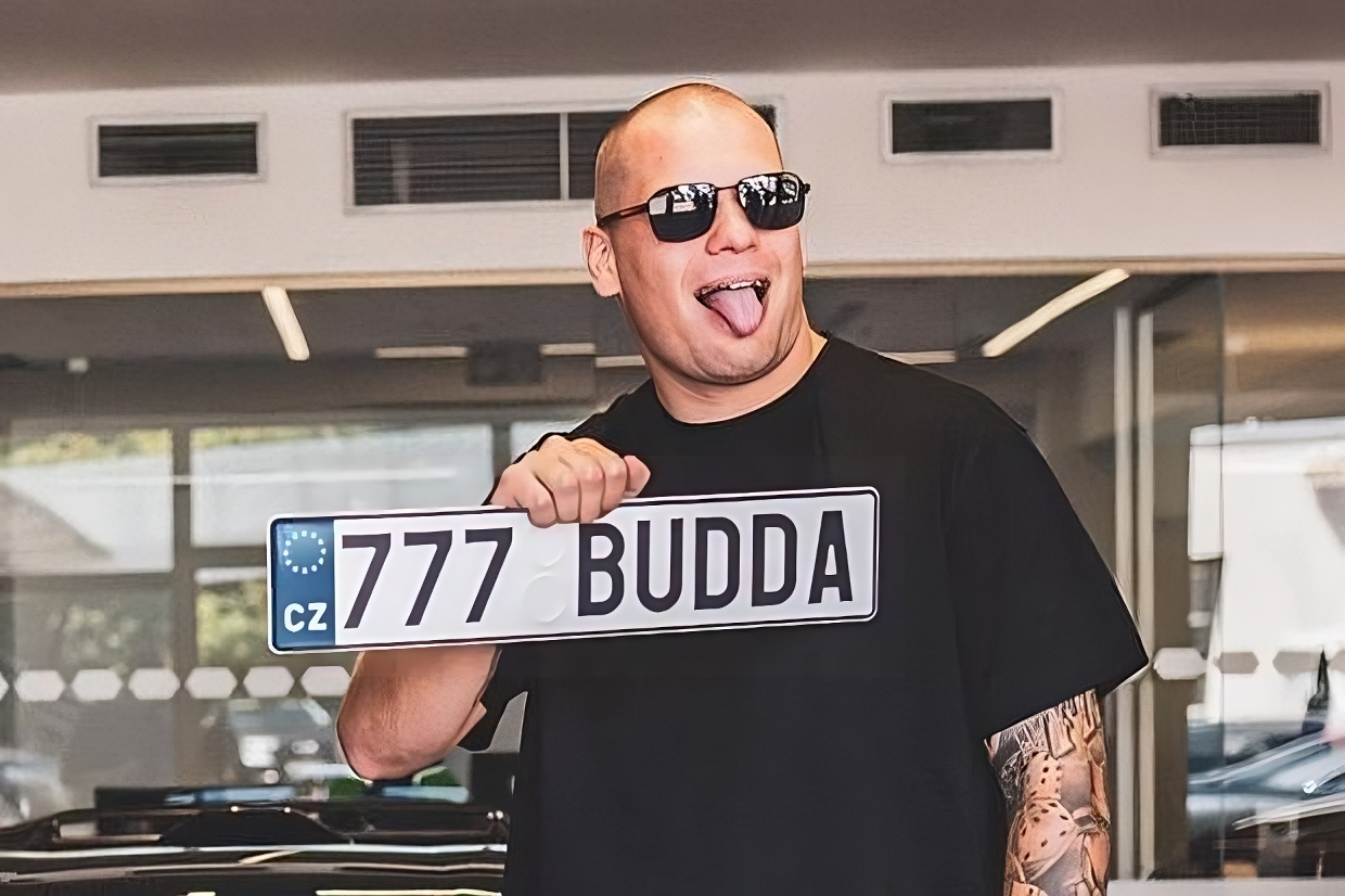YouTuber Budda w czarnych okularach, wystawiający język, trzymający tablicę rejestracyjną z napisem 777 Budda.