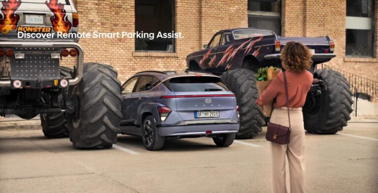 Kobieta korzysta z asystenta zdalnego parkowania, aby wyjechac Hyundaiem Kona II spomiędzy dwóch monster trucków